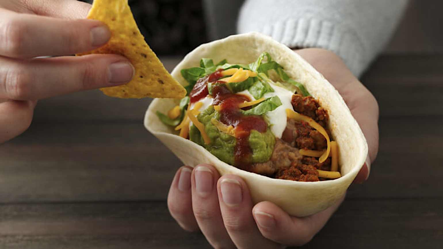 Une main tenant un bol de tortilla molle Old El Paso avec du guacamole, du bœuf haché, de la crème sure, du fromage, de la laitue et des haricots frits